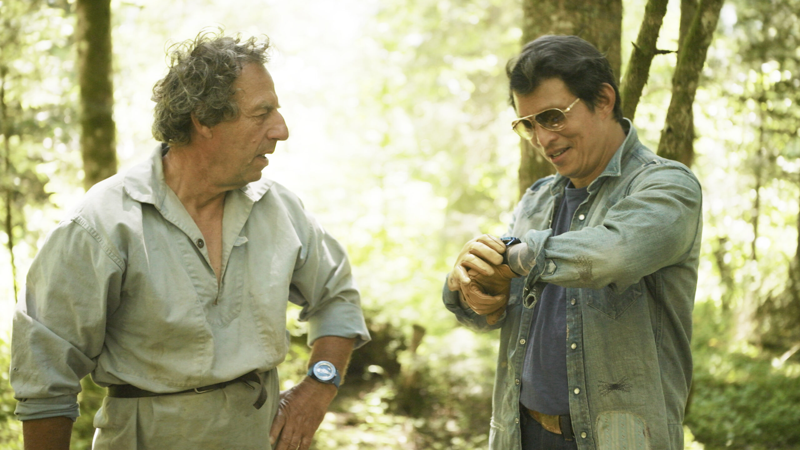 Wei Koh and Denis Flageollet standing in a sun-lit forest, talking while examining a watch during the Man of the Hour episode on De Bethune