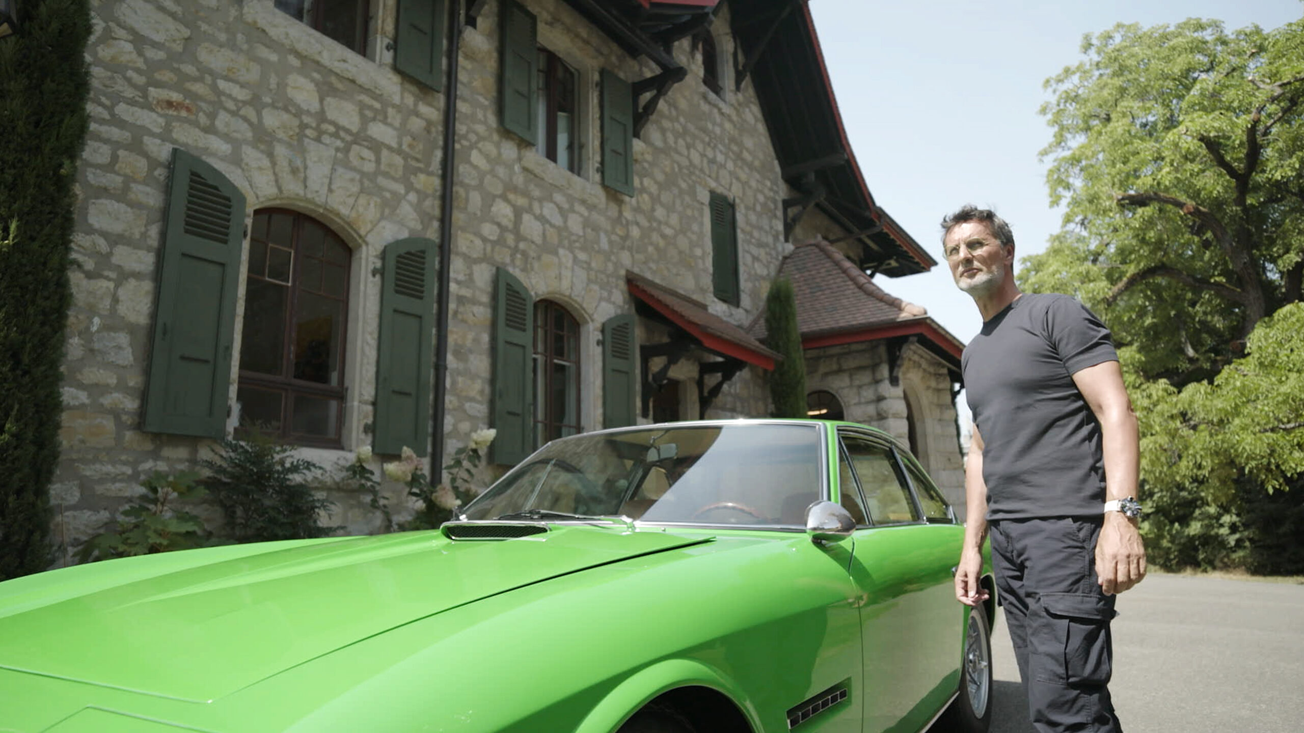 Max Büsser standing beside his bright green 1969 Lamborghini Islero outside his Geneva home, filmed for the Man of the Hour episode
